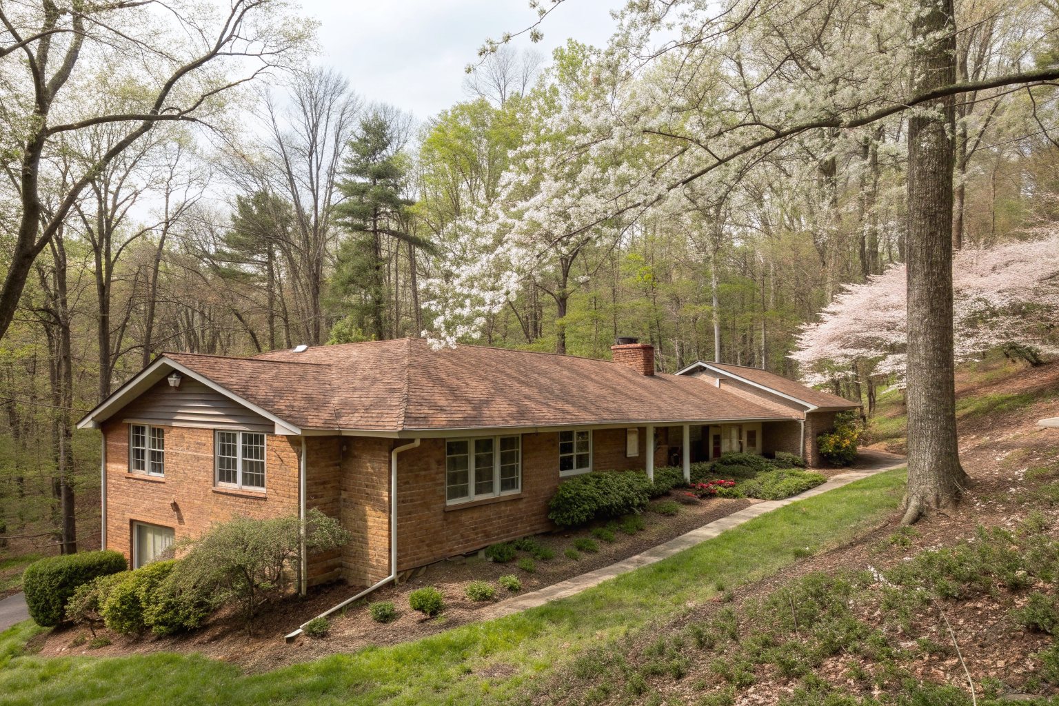 A 1960s brick ranch house with a faded brown shingle roof on a sloped wooded lot with flowering trees